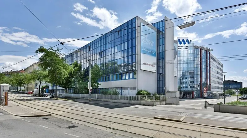 Modernes Bürogebäude mit markanter Glasfassade und Straßenbahnschienen im Vordergrund unter blauem Himmel.