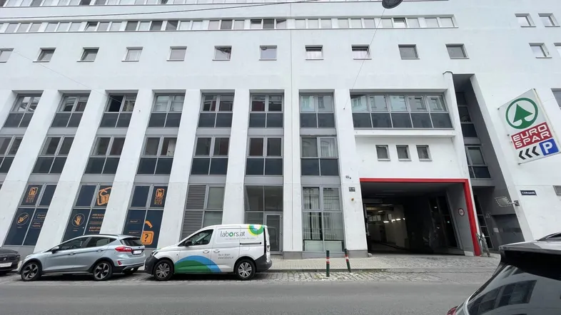 Modernes Gebäude mit vielen Fenstern, einer Tiefgarageneinfahrt und einem Supermarkt-Schild an der Fassade.