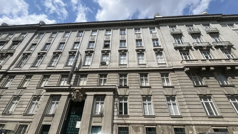 Graue Fassade eines Mehrfamilienhauses mit vielen Fenstern und kleinen Balkonen unter bewölktem Himmel.
