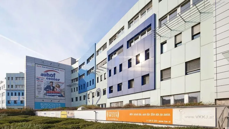 Moderne Bürogebäude-Fassade mit blauen Akzenten und großen Fenstern, umgeben von Grünflächen und Werbetafeln.