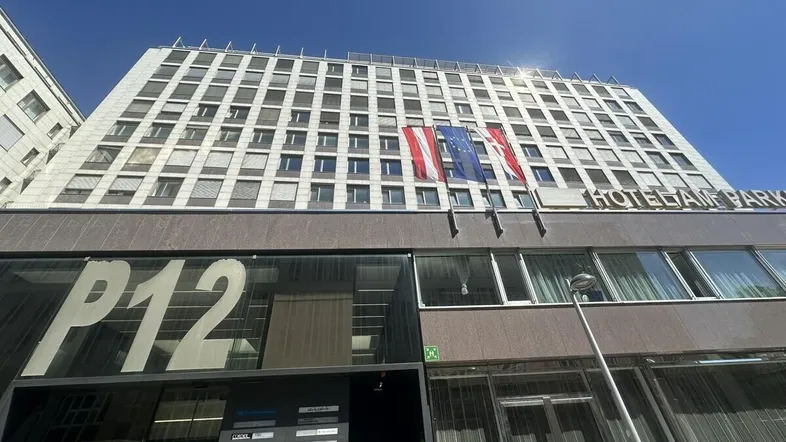 Gebäudefassade eines Hotels mit mehreren Stockwerken, großen Fenstern und gehissten Flaggen unter blauem Himmel.