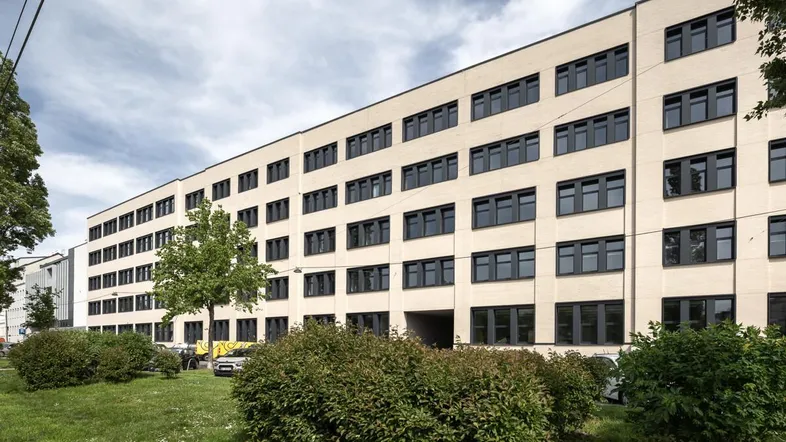 Modernes Bürogebäude mit vielen Fenstern und gepflegter Grünanlage davor unter bewölktem Himmel.