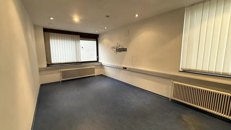 Büroraum mit blauen Vinylböden, mehreren Fenstern mit Jalousien und Wandheizkörpern.