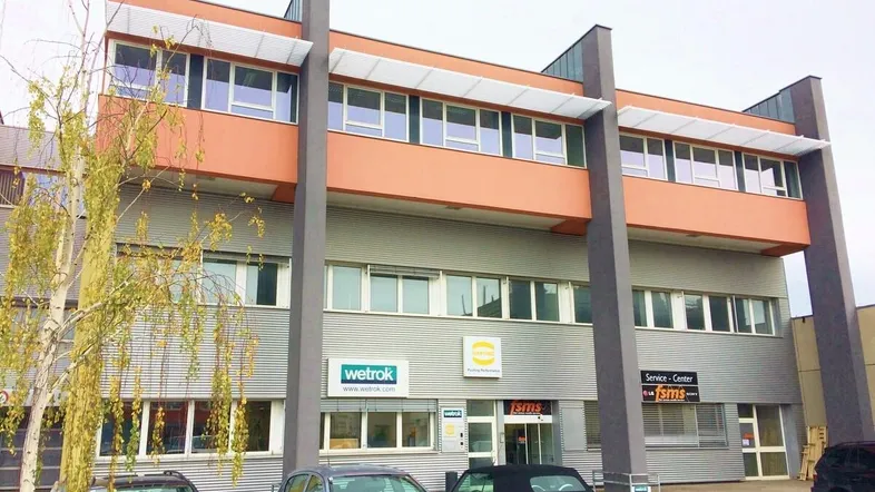 Modernes Geschäftsgebäude mit mehreren Etagen und Parkplätzen davor, unter einem bewölkten Himmel.