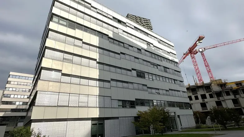 Modernes Bürogebäude mit vielen Fenstern und Jalousien, umgeben von Baustellenkränen unter bewölktem Himmel.