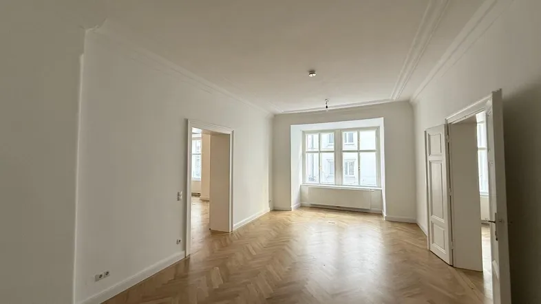 Geräumiges Wohnzimmer mit elegantem Fischgrätparkett, hohen Decken und großen Fenstern, die viel Tageslicht hereinlassen.