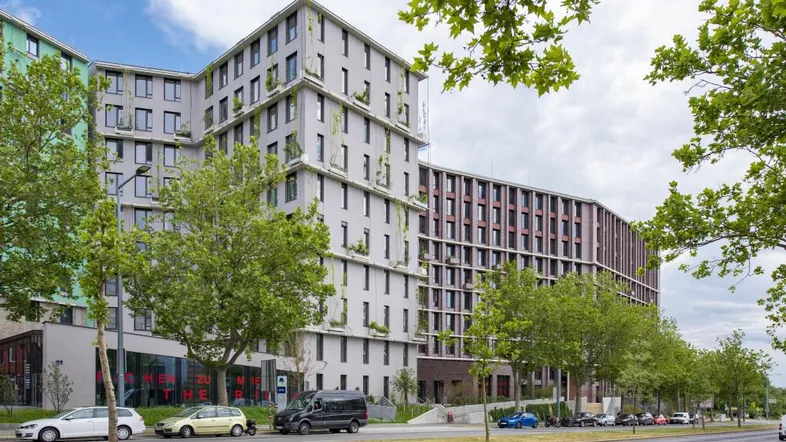 Moderne Wohngebäude mit begrünten Fassaden und einer belebten Straße davor unter bewölktem Himmel.