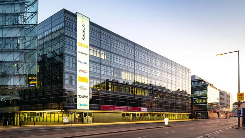 Modernes Bürogebäude mit Glasfassade und beleuchteten Werbetafeln an einer belebten Straße bei Sonnenuntergang.