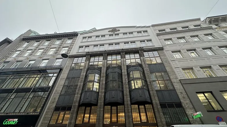 Historisches Gebäude mit markanter Fassade, großen Fenstern und Geschäften im Erdgeschoss in städtischer Umgebung.