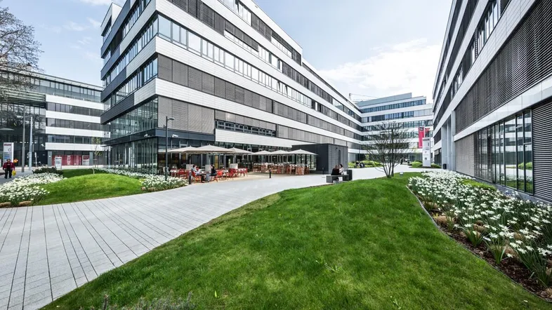 Moderne Bürogebäude mit gepflegten Grünflächen und Außengastronomie unter Sonnenschirmen.