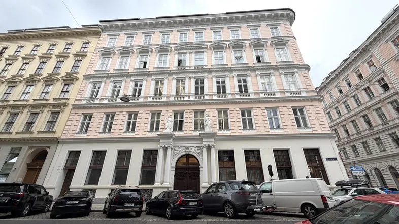 Historisches Gebäude mit rosa Fassade, vielen Fenstern und klassischer Architektur in einer Stadtstraße.