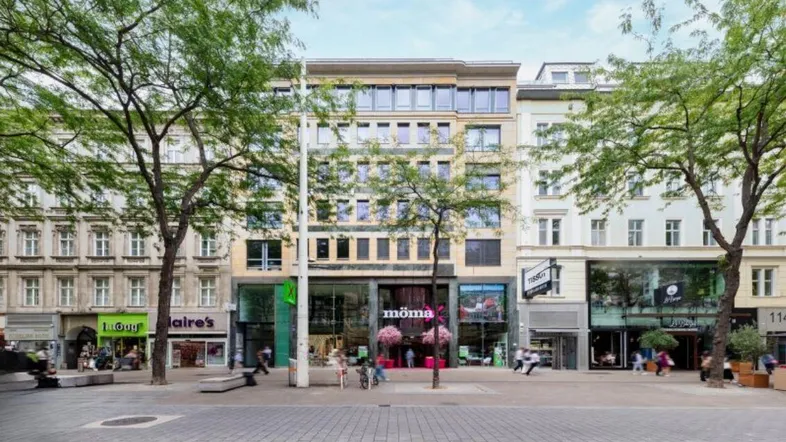 Eine belebte Einkaufsstraße mit Geschäften und Bäumen vor einem mehrstöckigen Gebäude mit moderner Architektur.