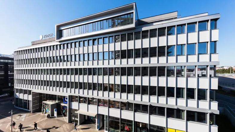 Modernes Bürogebäude mit vielen Fenstern und Geschäften im Erdgeschoss, umgeben von städtischer Infrastruktur.