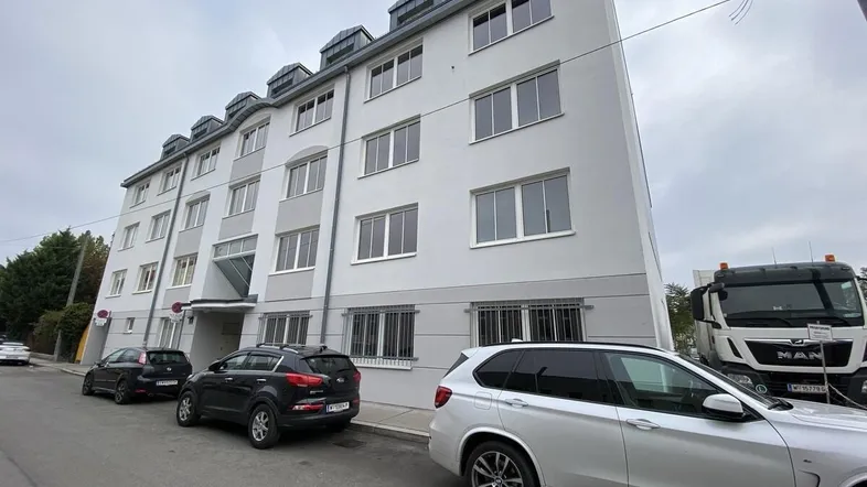 Mehrstöckiges Wohngebäude mit weißer Fassade und parkenden Autos auf der Straße unter bewölktem Himmel.