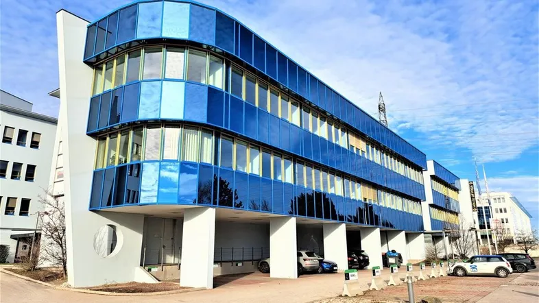 Modernes Bürogebäude mit auffälliger blauer Glasfassade und Parkplätzen davor unter blauem Himmel.