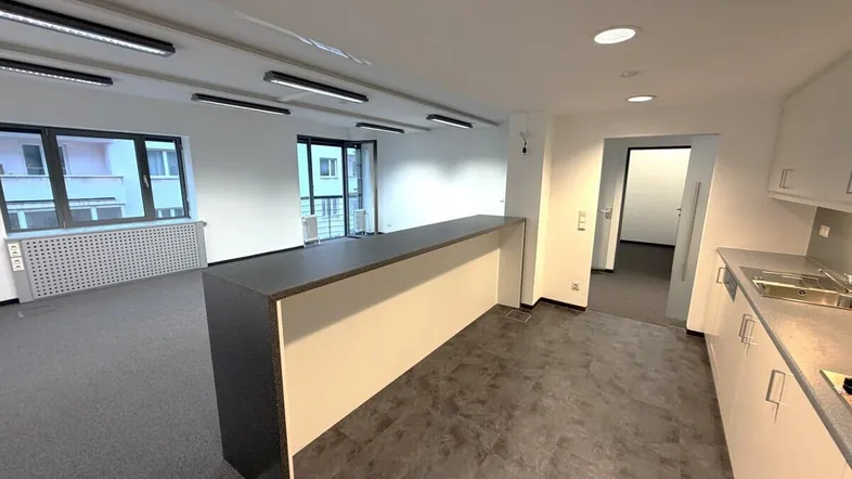 Modernes Büro mit integrierter Küchenzeile, Teppichboden und großen Fenstern, die viel Tageslicht hereinlassen.