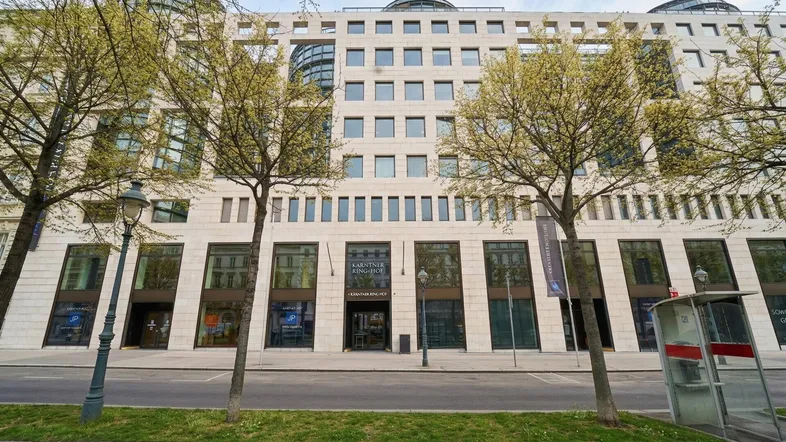 Fassade eines modernen Bürogebäudes mit großen Fenstern und Bäumen davor an einer belebten Straße.