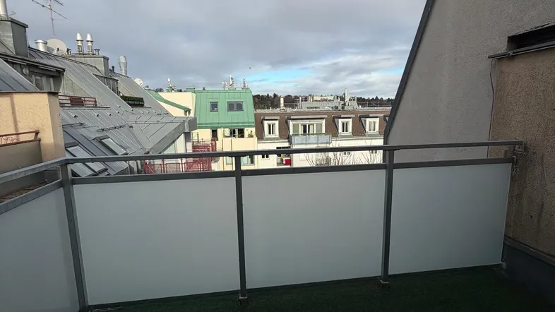 Geräumiger Balkon mit Kunstrasen und Blick über die Dächer der Stadt unter bewölktem Himmel.