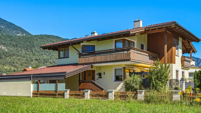 Traditionelles Haus mit Holzbalkonen und Satteldach, umgeben von grüner Landschaft und Bergen.
