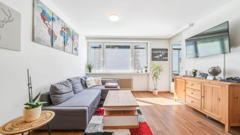 Helles Wohnzimmer mit gemütlichem Sofa, Holzboden und Blick nach draußen durch das Fenster.