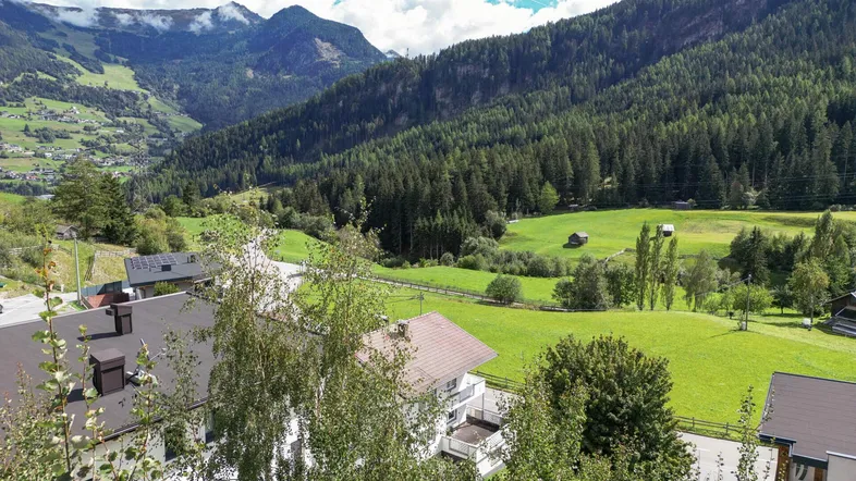 Panoramablick über ein grünes Tal mit Häusern, Wäldern und majestätischen Bergen unter blauem Himmel.