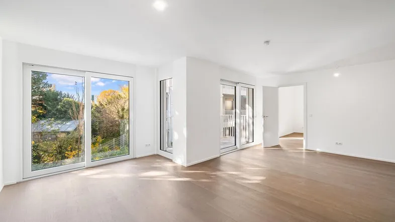 Heller, leerer Raum mit großen Fenstern, Holzboden und Zugang zu einem Balkon.