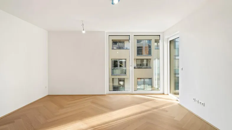 Helles, leeres Zimmer mit Fischgrätparkett und großen Fenstern, die viel Tageslicht hereinlassen.