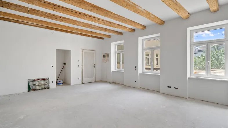 Geräumiger Wohnbereich im Rohbau mit sichtbaren Holzbalken und hellen Fenstern.