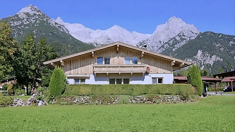 Traditionelles Haus mit Holzbalkon und beeindruckendem Bergblick, umgeben von grüner Landschaft.