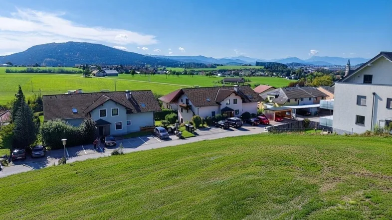 Luftaufnahme einer ländlichen Wohnsiedlung mit grünen Feldern und Bergen im Hintergrund unter blauem Himmel.