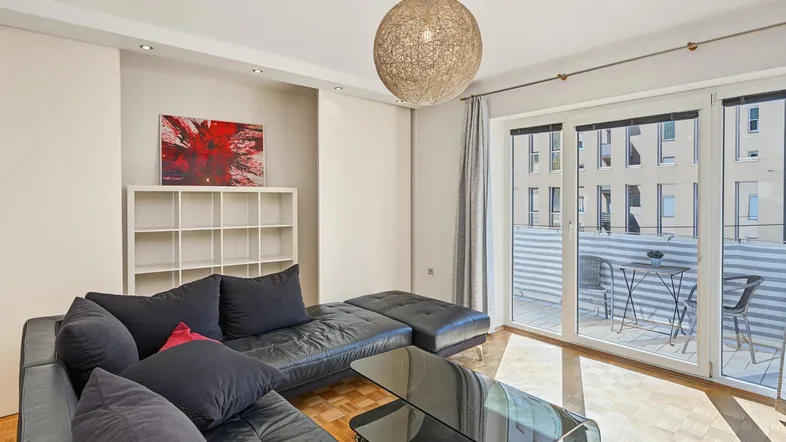 Helles Wohnzimmer mit dunklem Sofa, Couchtisch und Zugang zu einem Balkon.
