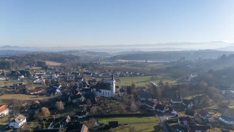 Luftaufnahme eines malerischen Dorfes mit Kirche, umgeben von Feldern und sanften Hügeln unter blauem Himmel.
