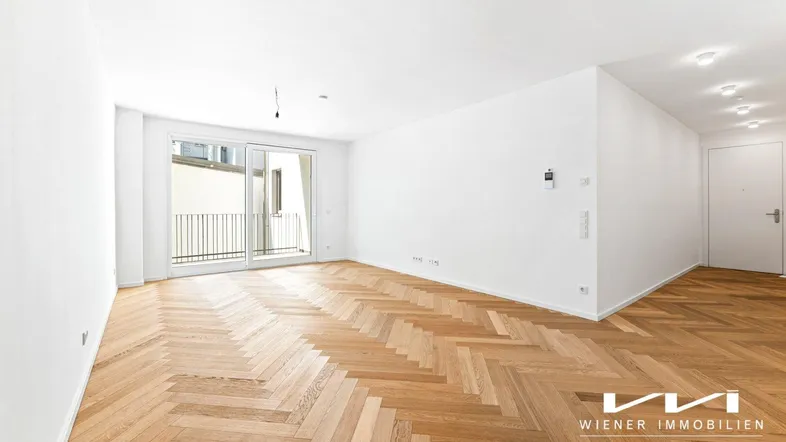 Helles, leeres Wohnzimmer mit Fischgrätparkett und Zugang zu einem Balkon.