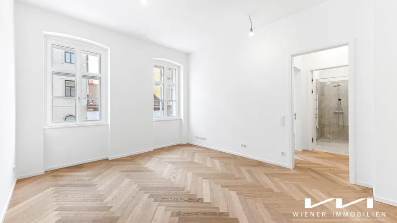 Helles, leeres Zimmer mit Fischgrätparkett und zwei großen Fenstern, das viel Tageslicht hereinlässt.