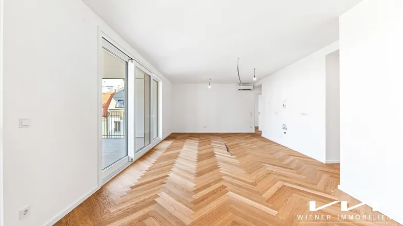 Helles, leeres Wohnzimmer mit Fischgrätparkettboden und Zugang zu einem Balkon oder einer Terrasse.