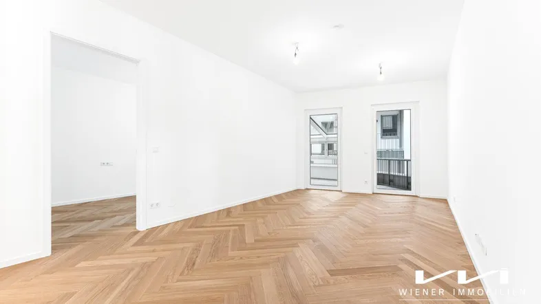Helles, leeres Zimmer mit Fischgrätparkettboden und Zugang zu einem französischen Balkon.