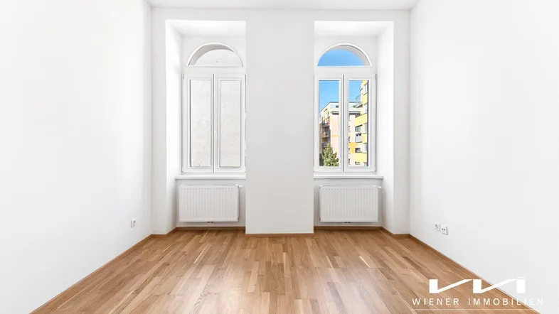 Helles Zimmer mit Parkettboden und zwei hohen Fenstern, die viel Tageslicht hereinlassen.