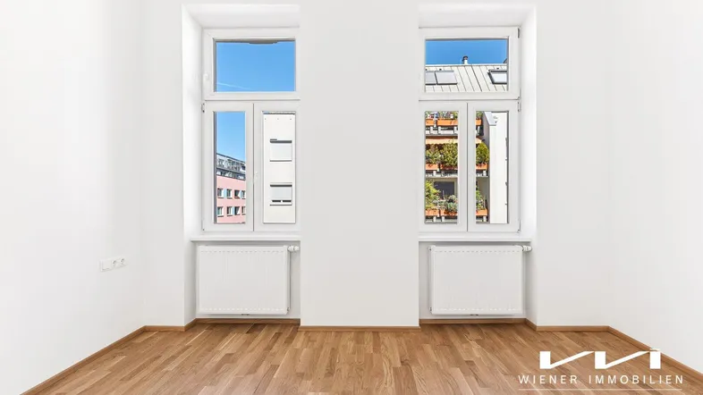 Helles Zimmer mit Parkettboden und zwei großen Fenstern, die viel Tageslicht hereinlassen.