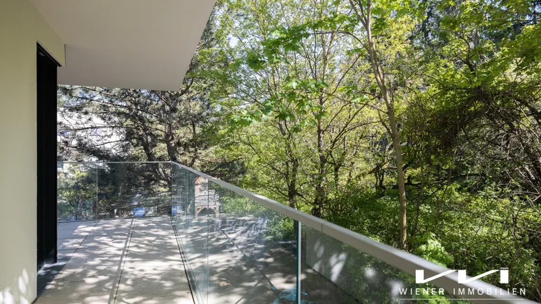 Geräumiger Balkon mit Glasgeländer und Blick ins Grüne, umgeben von Bäumen und viel Tageslicht.