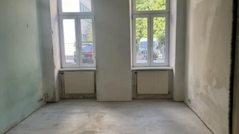 Leerer Raum in einem Altbau mit zwei großen Fenstern und Betonboden, bereit für die Renovierung.