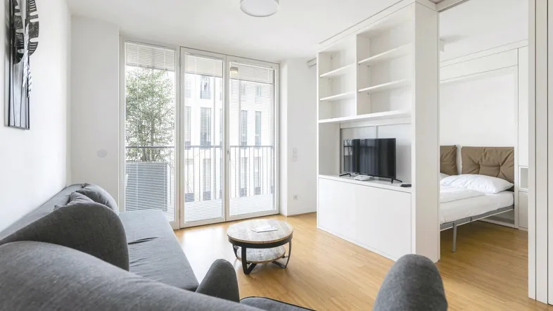 Helles Wohnzimmer mit Holzboden, integriertem Regal und Zugang zu einem kleinen Balkon.