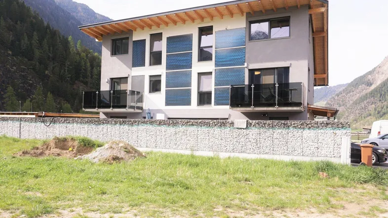 Modernes Mehrfamilienhaus mit Solarpaneelen an der Fassade und Bergblick in ländlicher Umgebung.