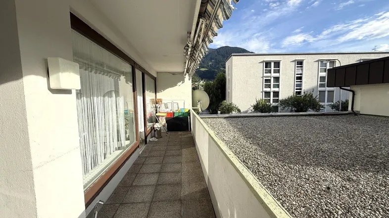 Langer Balkon mit Fliesenboden, Markise und Blick auf umliegende Gebäude und Berge.
