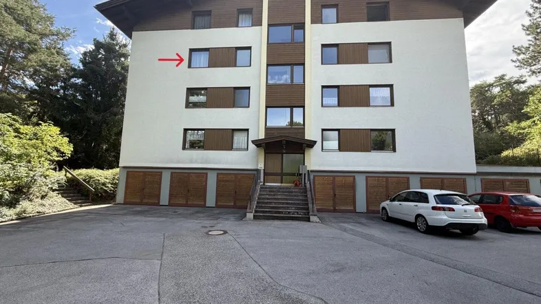 Mehrfamilienhaus mit mehreren Etagen, Garagen und Parkplätzen, umgeben von Natur.
