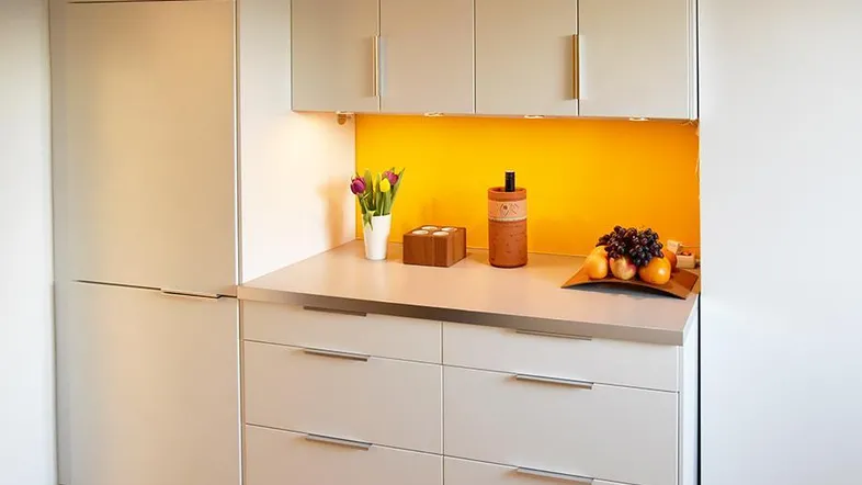 Moderne Küchenzeile mit beigen Schränken, Edelstahl-Arbeitsplatte und leuchtend gelber Rückwand.