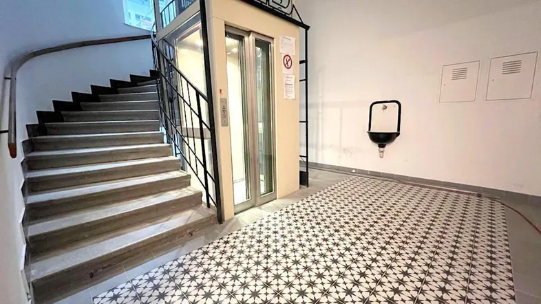 Heller Eingangsbereich mit modernem Aufzug, Treppe und gemusterten Bodenfliesen.