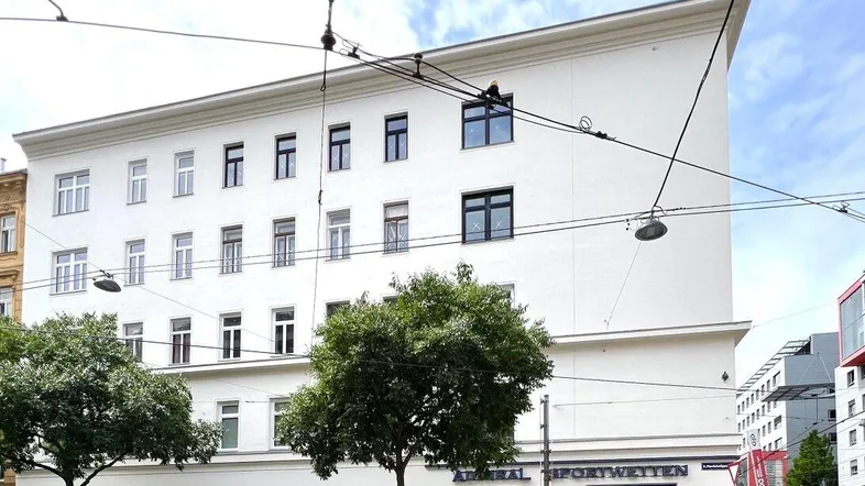 Weißes Mehrfamilienhaus mit vielen Fenstern und Geschäften im Erdgeschoss an einer belebten Straße.