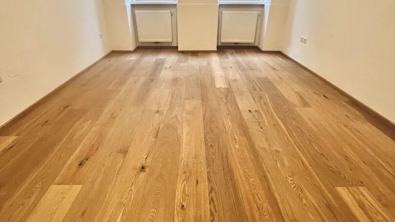 Helles Zimmer mit neuem Holzparkettboden und zwei Fenstern, die auf die Straße blicken.