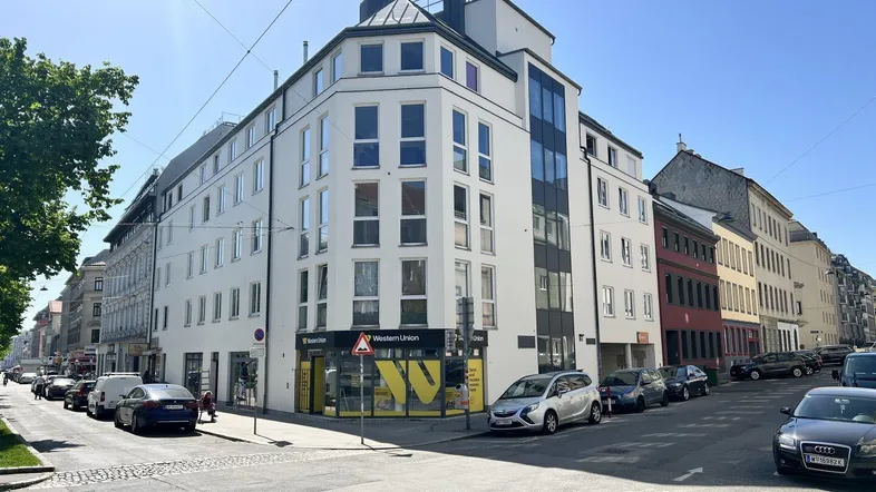 Modernes Eckgebäude an einer belebten Straße mit Geschäften im Erdgeschoss und vielen Fenstern.