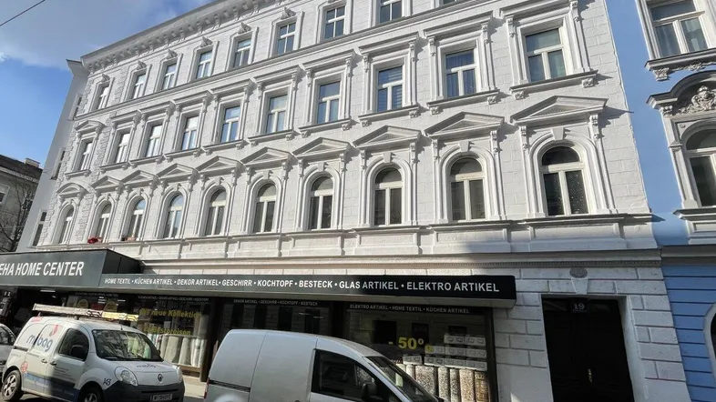 Klassisches Mehrfamilienhaus mit weißer Fassade und vielen Fenstern an einer belebten Straße.